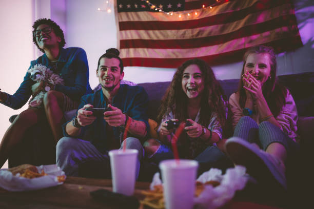 Young multi-ethnic friends eating fast food and playing video games Multi-ethnic friends hanging out on living room sofa eating fast food and playing video games college dorm party stock pictures, royalty-free photos & images