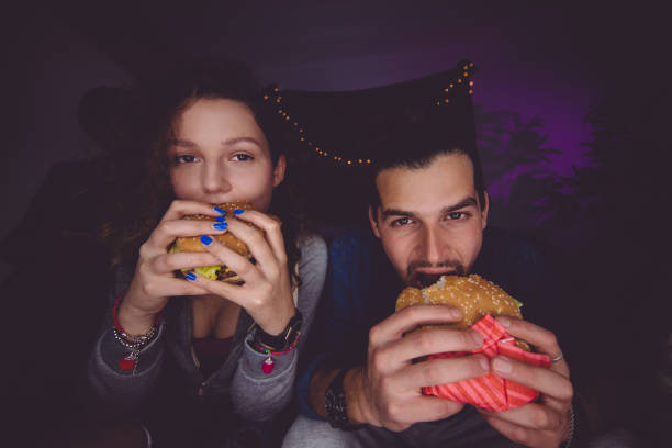 Portrait of young man and woman eating hamburgers Portrait of young multi-ethnic friends having unhealthy diet, eating fast food and biting hamburgers college dorm party stock pictures, royalty-free photos & images