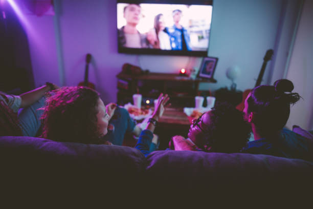 Multi-ethnic teenage friends watching TV together at hangout house Young multi-ethnic hipster friends relaxing on living room sofa and watching film on TV watching tv stock pictures, royalty-free photos & images