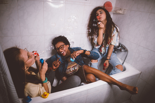 Young multi-ethnic hipster girls having slumber party at home and blowing bubbles in bathtub
