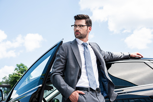 portrait of thoughtful businessman in eyeglasses looking away while standing at car on street