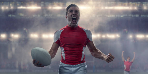 Close up of a professional male rugby player holding rugby ball, shouting with clenched fist in celebration of a victory. The player is standing in a generic floodlit rugby stadium full of spectators during a rugby match in cold and rainy conditions. The player wears generic red and white kit.