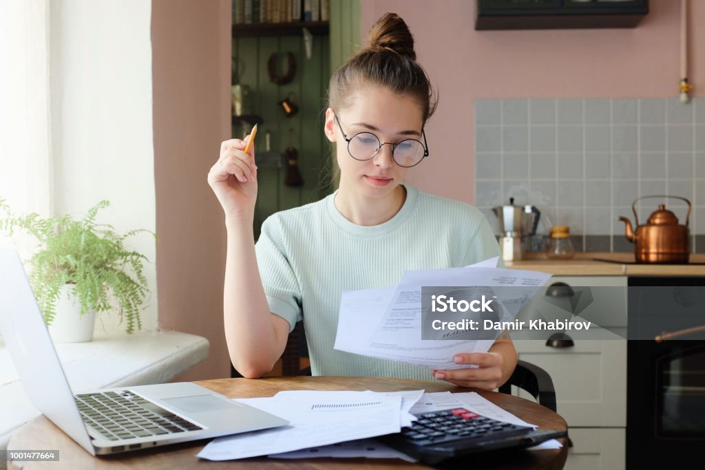 Indoor picture of young European female sitting at home at table Indoor picture of young European female sitting at home at table reading data in sheets of paper with pencil in order to figure out something important and check it, feeling relaxed and confident Financial Bill Stock Photo