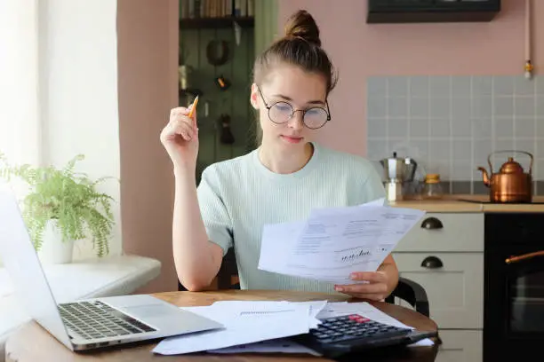 Indoor picture of young European female sitting at home at table reading data in sheets of paper with pencil in order to figure out something important and check it, feeling relaxed and confident