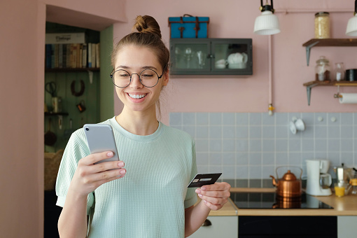 Closeup photo of European Caucasian girl using mobile phone and credit card to pay for goods and services online, looking satisfied because of saved time and effort, enjoying leisure time at home