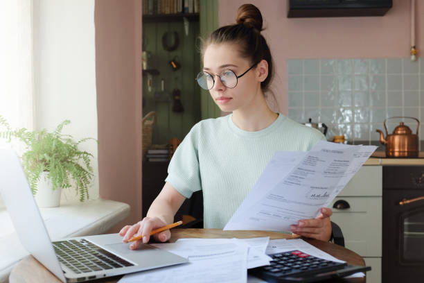 молодая женщина, управляющая домашним бюджетом, сидя за кухонным столом с открытым ноутбуком, документами и калькулятором, используя сенсо - loan house calculator currency стоковые фото и изображения
