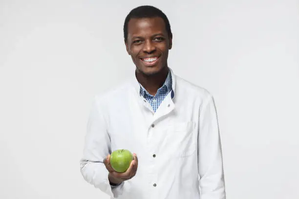 Photo of Indoor picture of young African American dentist standing in white uniform against white background with green apple in hand symbolizing necessity of taking care of teeth, smiling positively
