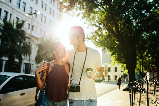 Happy explorer couple walking in the street holding a coffee cup with sun in the background. Man carrying a camera and holding a coffee cup walking with a woman with his arm around her shoulders.