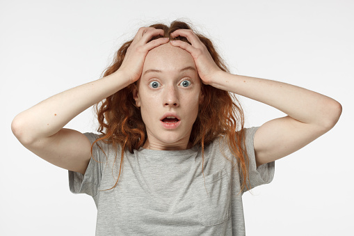 Headshot of handsome surprised redhead female looking at the camera, astonished, holding hands on head, standing against grey background. Human face expression concept