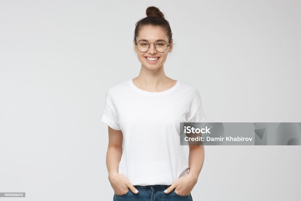 Joven Europea de pie con las manos en los bolsillos, vistiendo la camiseta blanca en blanco con espacio de copia de su logotipo o texto, aislado en fondo gris - Foto de stock de Camiseta libre de derechos