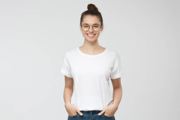 joven europea de pie con las manos en los bolsillos, vistiendo la camiseta blanca en blanco con espacio de copia de su logotipo o texto, aislado en fondo gris - glasses women smiling human face fotografías e imágenes de stock