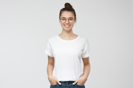 Young european woman standing with hands in pockets, wearing blank white tshirt with copy space for your logo or text, isolated on grey background