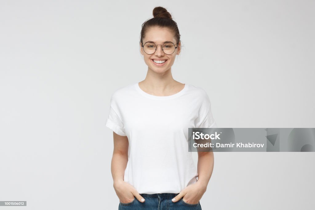 Young european woman standing with hands in pockets, wearing blank white tshirt with copy space for your logo or text, isolated on grey background T-Shirt Stock Photo