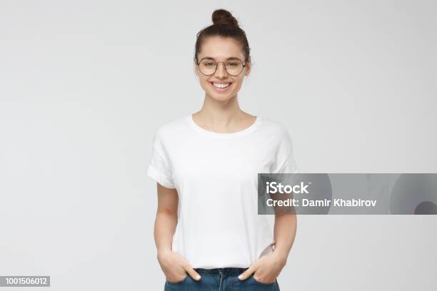 Young European Woman Standing With Hands In Pockets Wearing Blank White Tshirt With Copy Space For Your Logo Or Text Isolated On Grey Background Stock Photo - Download Image Now