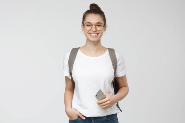 молодая женщина в белой футболке и траспарентных очках изолирована на сером фоне, улыбается, стоя в руках в карманах позирует - letter t фотографии стоковые фото и изображения