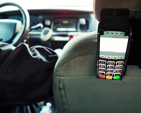 Horizontal image of a card reader attached to the back seat of a taxi, with front interior of vehicle and driver out of focus in the background, there is copy space.
