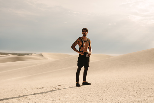 Full length of young muscular man standing on sand dune and looking away with hands on his hips. Fit man in shorts and water bladder pack workout in desert.