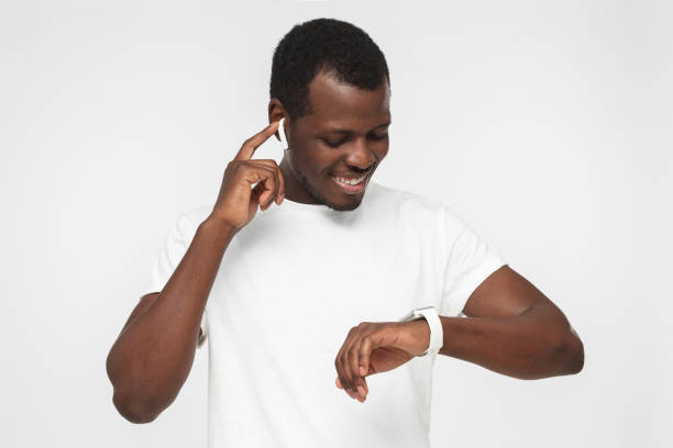 closeup fotografia de jovens afro-americanos homem retratado isolado no fundo cinza, olhando atentamente com sorriso positivo para tela do seu relógio digital no pulso ouvir música através de fones de ouvido - shorthair cat audio - fotografias e filmes do acervo