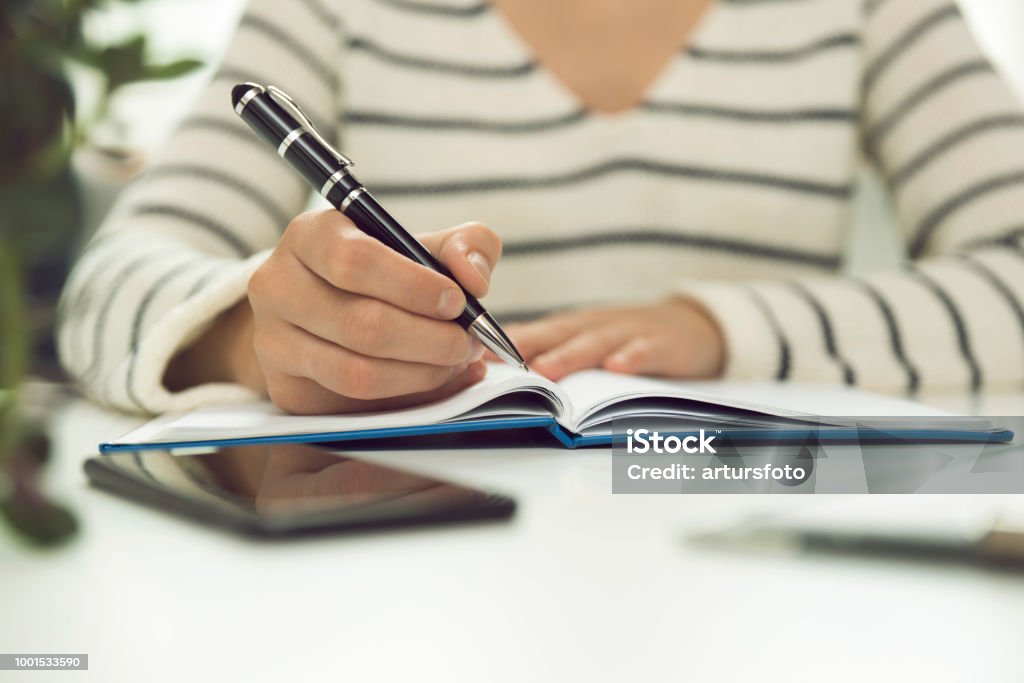 Young business woman sitting at table and writing in notebook. On table is smartphone, and tablet. Freelancer working, writing down new ideas Adult Stock Photo