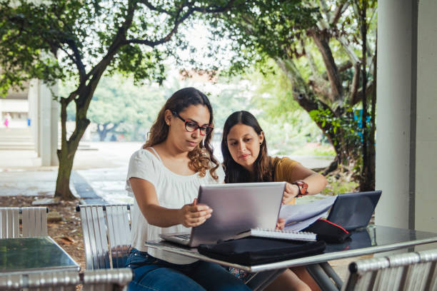 Two Hispanic Female Students, Collaborating , They Are Seated Outdoors Working On Laptops Girls sat at an outdoor desk working together on laptops, these two Hispanic university students are communicating and assisting one another, in the background the campus is blurred, with unidentifiable buildings and greenery, these friends are real people. valle del cauca stock pictures, royalty-free photos & images