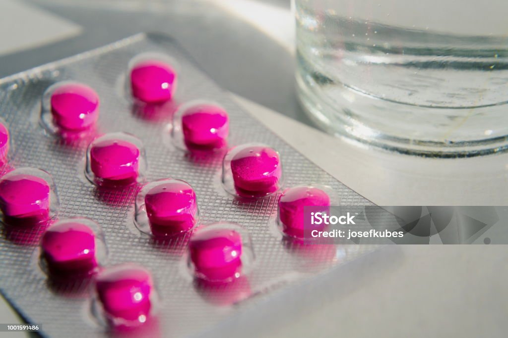 Pink pills with glass of water isolated on white background Detail of bright pink pills in medicine capsule with glass of water isolated on white background, pain migraine fever illness medicine concept Ibuprofen Stock Photo
