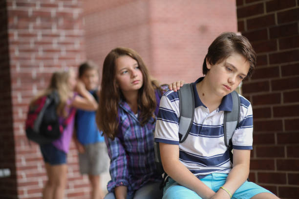 Comforting friend after being bullied. Friend comforts fellow student after he has been bullied outside the elementary school building,  Other students in background laugh at boy behind his back. school exclusion stock pictures, royalty-free photos & images