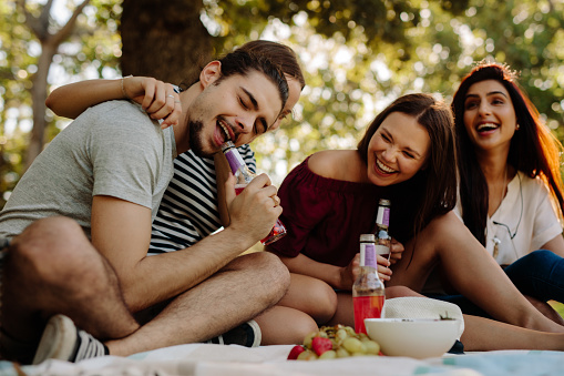 Man opening a bottle with his teeth and women sitting by smiling. Group of friends enjoying with drinks at picnic.