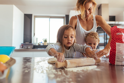 Mother and little children making cookies in kitchen. Siblings with rolling pin and flour helping mother in kitchen at home