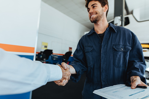 Young auto mechanic shaking hands with satisfied customer in garage. Automobile service center worker shaking hands with client after car servicing.