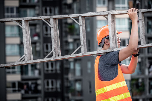 Trabajador de mantenimiento asiática joven con casco de seguridad naranja y chaleco con escalera de aluminio en obra. Ingeniería civil, constructor de arquitectura y construcción de conceptos de servicio photo
