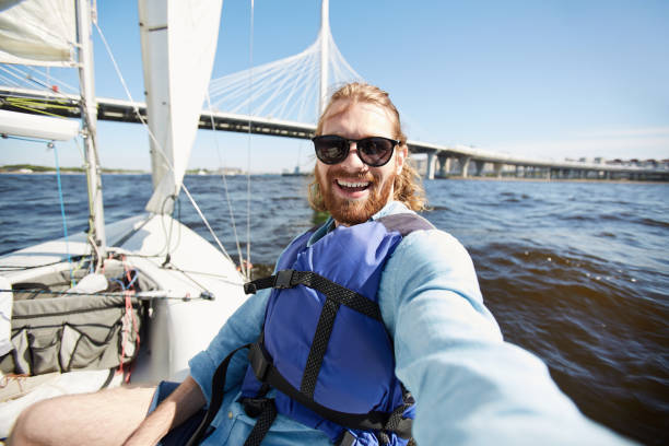 Excited man photographing himself on yacht Happy excited handsome young man with beard wearing sunglasses laughing while photographing himself against urban bridge while floating on yacht yacht selfie stock pictures, royalty-free photos & images