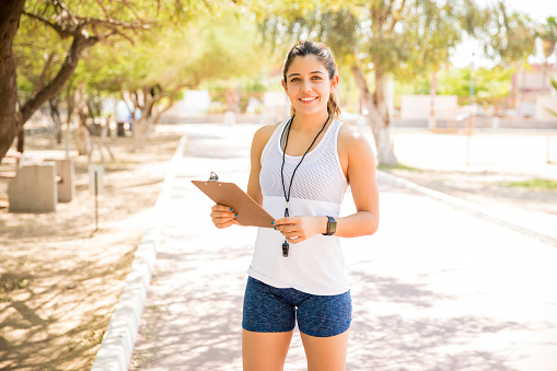Portrait of an attractive female gym coach taking some notes and smiling at camera while standing outdoors at park