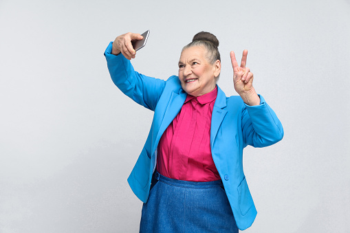 aged woman making selfie with peace sing and smilling. Portrait of handsome expressive grandmother in light blue suit with collected gray hair bun hairstyle. Studio shot, isolated on gray background