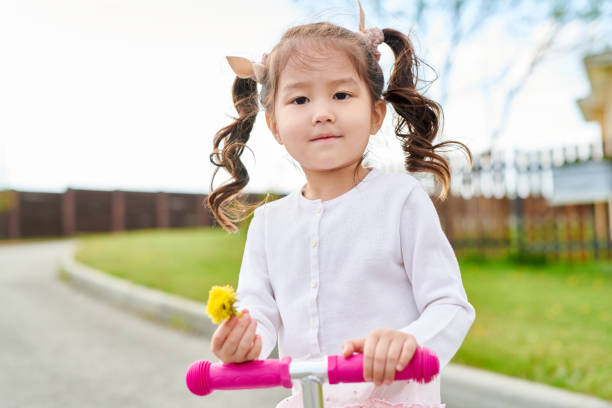 Waist up portrait of cute Asian girl riding scooter looking at camera while going down road and holding flower, copy space