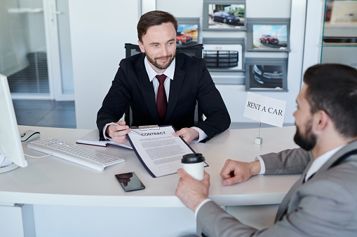 Confident salesman in suit working with client at the table. He is showing the terms of the contract of rent a car