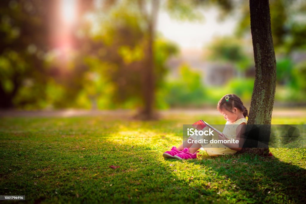 A little cute girl in a yellow dress reading a book sitting under the tree Child Stock Photo