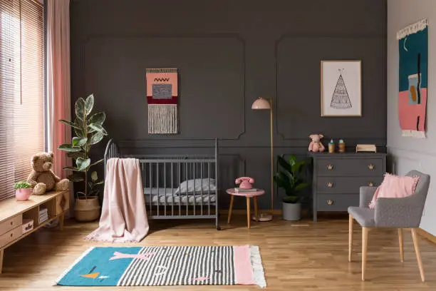 Photo of Real photo of a grey crib standing next to a pink stool, a lamp and cupboard in grey baby room interior also with armchair, rug and posters