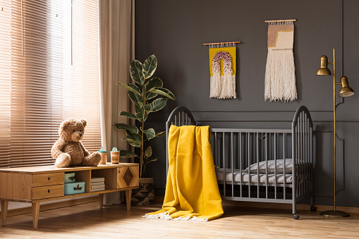 Foto real de una cuna con un pie de manta amarilla entre un armario bajo con un oso y una lámpara en el interior de habitación de bebé photo
