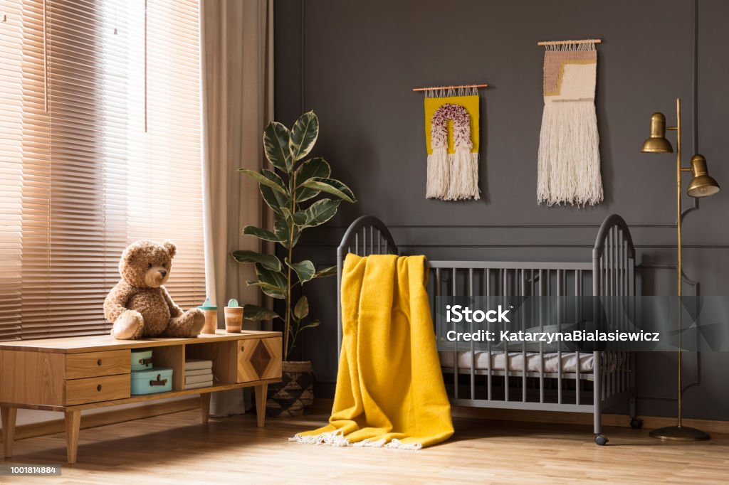Echte foto van een kinderbed met een gele deken staan tussen een lage kast met een beer en een lamp in baby kamer interieur - Royalty-free Kinderkamer Stockfoto