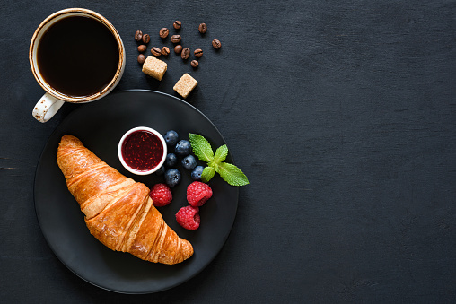 Croissant with berries, jam and cup of black coffee on black background. Top view with copy space. Breakfast concept