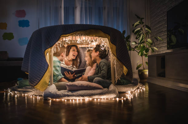 young cheerful nanny reading stories to small kids in a tent at home. - family reading african descent book imagens e fotografias de stock