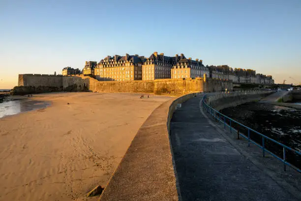 Photo of View at sunset of the walled old city of Saint-Malo in Brittany, France, with granite buildings bathed in warm light protruding above the wall and the Mole beach at the foot of the ramparts.