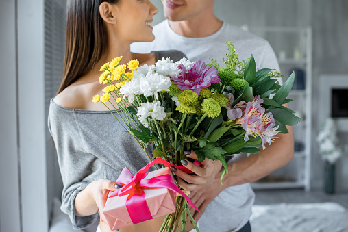 partial view of man hugging smiling woman with present and bouquet of flowers at home