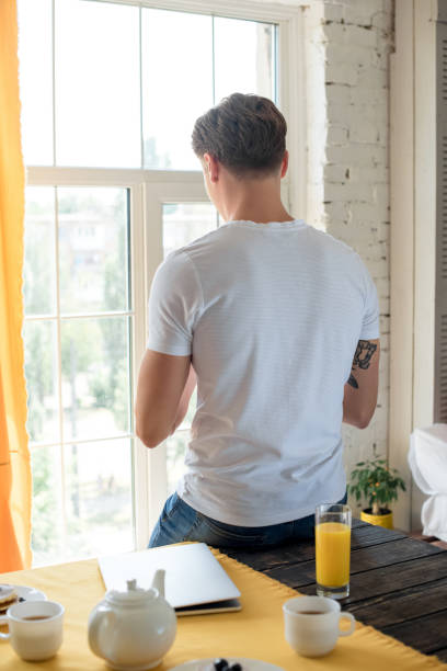 back view of man standing at window with breakfast on table at home