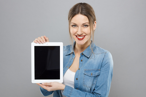 Indoor portrait of beautiful pleased European woman isolated on grey background presenting blank tablet device with wide happy smile with copyspace for advertising applications, products and services.