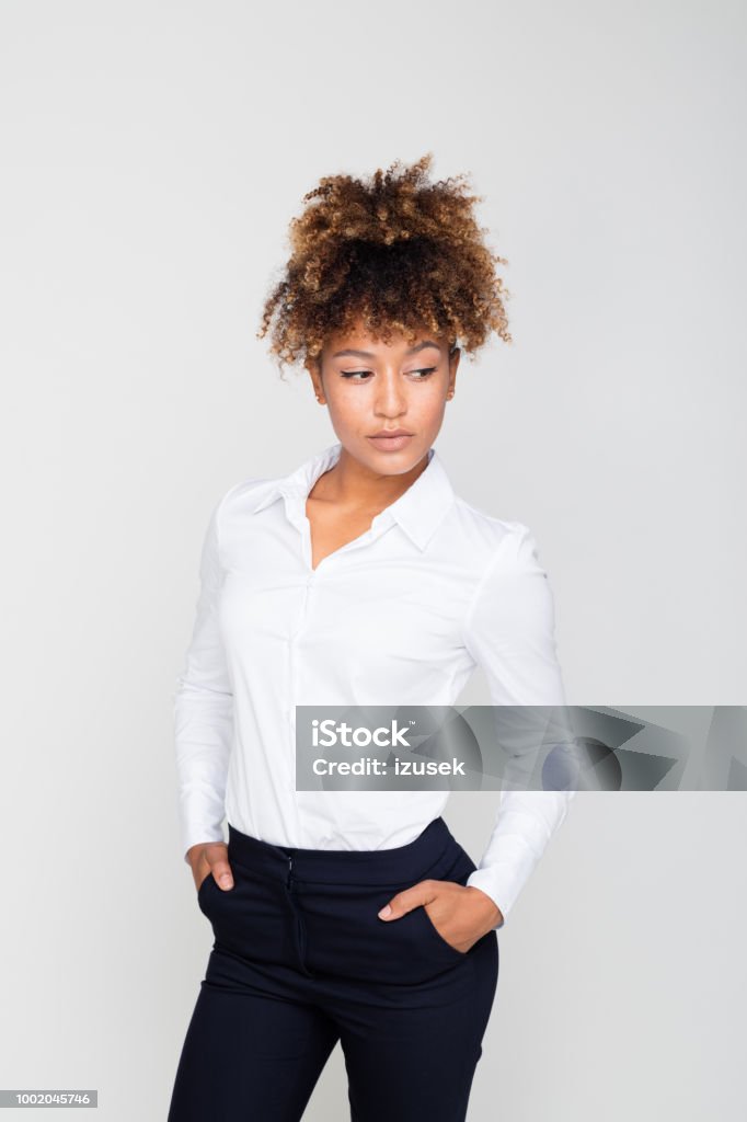 Portrait of pensive afro amercian businesswoman Thoughtful afro american businesswoman wearing white shirt standing with hands in pockets against grey background. Studio shot. Adult Stock Photo