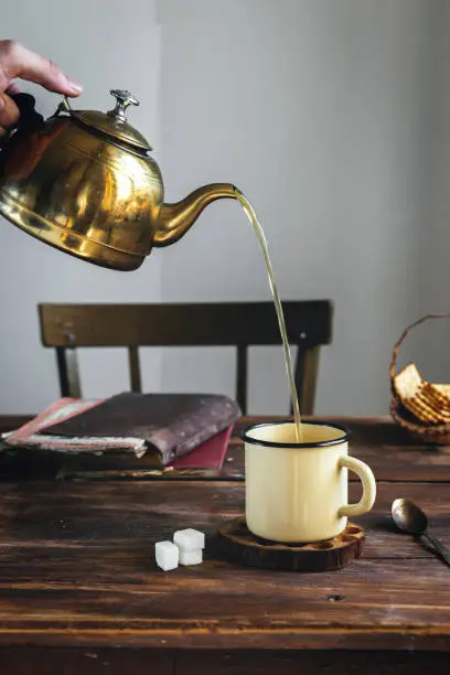 Rustic style. Pouring hot tea in a metal mug, a metal teapot, sugar cubes, cookies, on an old wooden table