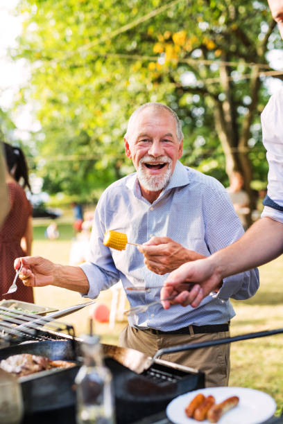 A senior man standing by the grill, holding corn on a barbecue party.