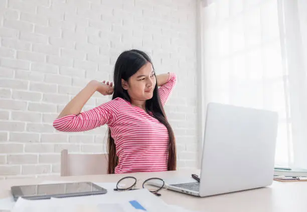 Asian woman freelancer stretching arm at back in front of laptop on desk from work in home office,work at home