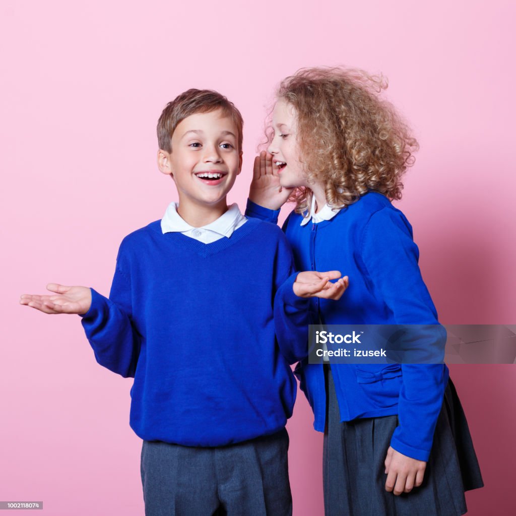 Portrait of cute and happy schoolboy and schoolgirl Schoolgirl whispering to her cute school friend. Studio shot, pink background. Child Stock Photo